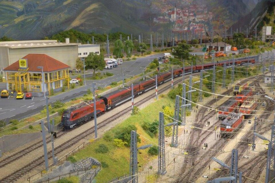 Ein Modellbahnansicht mit mehreren Zügen auf Schienen. Im Hintergrund sind Gebäude und eine Straße mit Autos zu sehen. | © Modeleisenbahn Knittelfeld