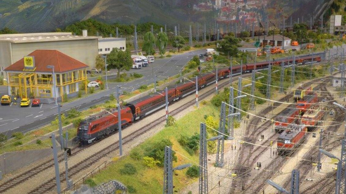 Ein Modellbahnansicht mit mehreren Zügen auf Schienen. Im Hintergrund sind Gebäude und eine Straße mit Autos zu sehen. | © Modeleisenbahn Knittelfeld