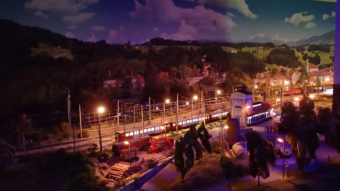 Eine detaillierte Modelleisenbahn mit beleuchteten Zügen und einer malerischen Landschaft. Im Hintergrund sind Berge und eine Nachtansicht mit warmem Licht zu sehen. | © Modeleisenbahn Knittelfeld