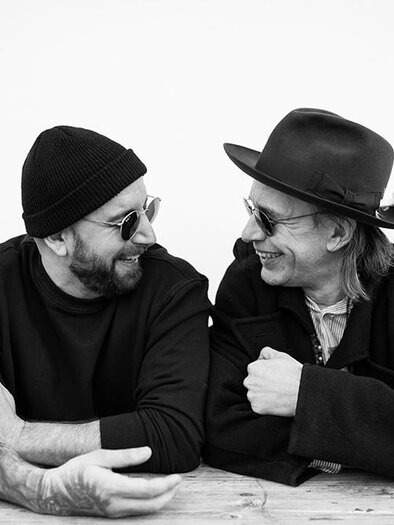 Two men are laughing and having a cheerful conversation at a wooden table. They are wearing sunglasses and hats and seem to be having a great time. | ©  Daniela Matejschek