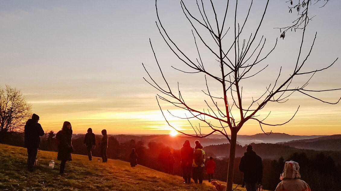 Eine Gruppe von Menschen steht auf einer Wiese und schaut auf einen wunderschönen Sonnenuntergang. Im Hintergrund sind sanfte Hügel und ein klarer Himmel zu sehen.