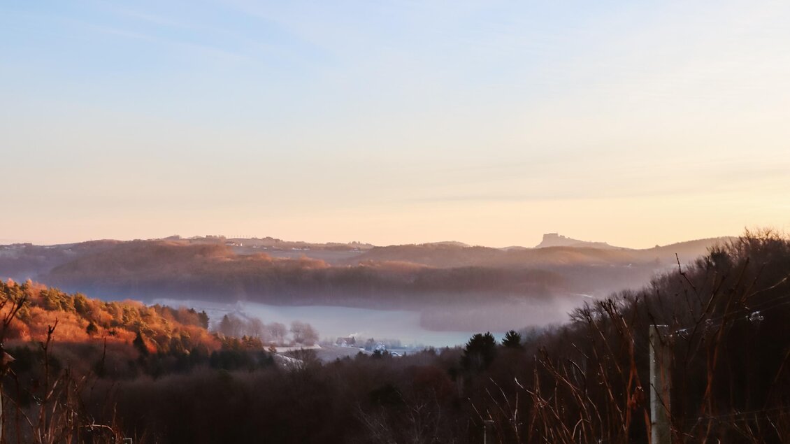Eine malerische Landschaft mit sanften Hügeln und wassergefüllten Tälern in der Morgendämmerung. Der Himmel zeigt warme Farbtöne und es liegt Nebel über dem Tal.