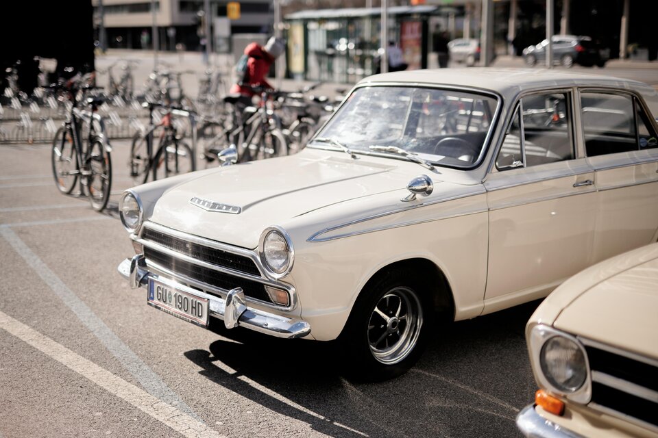 Ein klassisches, weißes Auto steht auf einem Parkplatz. Im Hintergrund sind mehrere Fahrräder abgestellt.