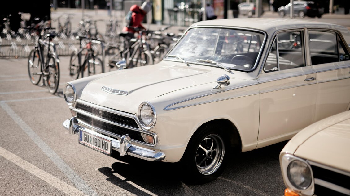 Ein klassisches, weißes Auto steht auf einem Parkplatz. Im Hintergrund sind mehrere Fahrräder abgestellt.