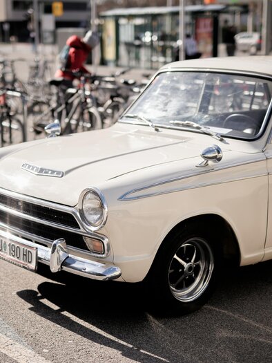 Ein klassisches, weißes Auto steht auf einem Parkplatz. Im Hintergrund sind mehrere Fahrräder abgestellt.
