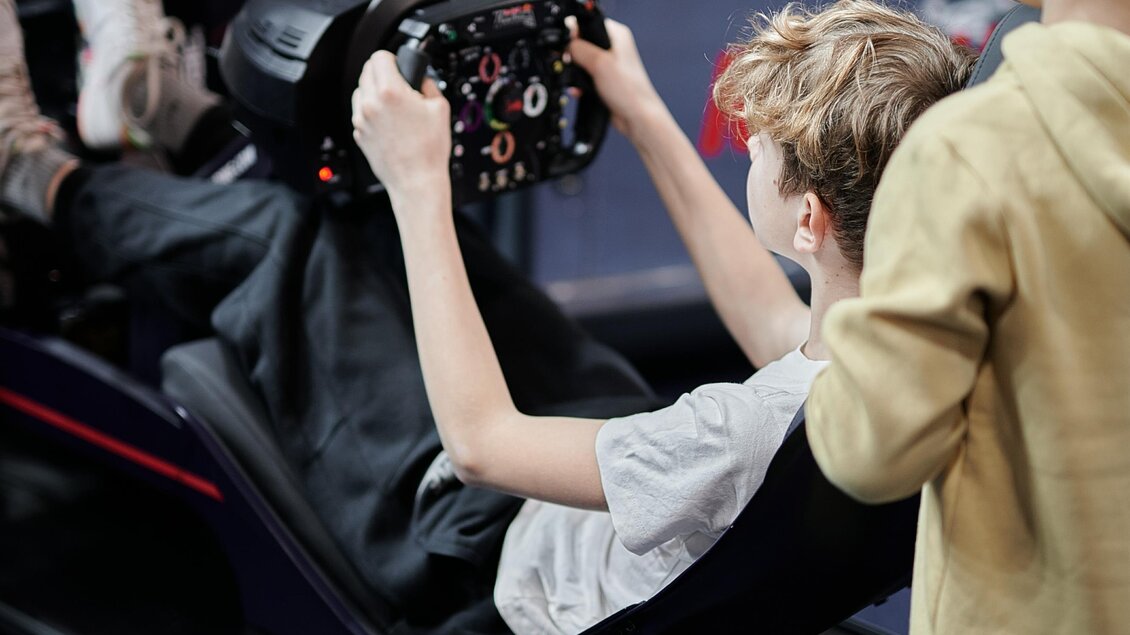 Ein Junge sitzt in einem Rennsimulator und steuert mit einem Lenkrad. Ein weiterer Junge schaut interessiert zu. | © Valerie_Maltseva