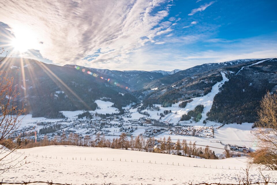 Eine malerische Winterlandschaft mit schneebedeckten Bergen und einer kleinen Stadt im Tal. Die Sonne scheint hell am Himmel und schafft eine gemütliche Atmosphäre. | © Tv Murau