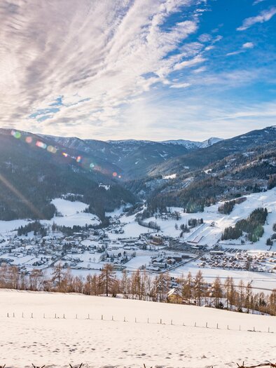 A picturesque winter landscape with snow-covered mountains and a small town in the valley. The sun shines brightly in the sky, creating a cozy atmosphere. | © Tv Murau