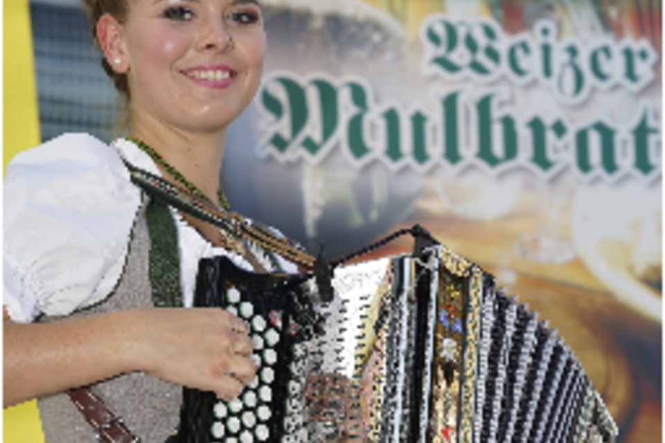 Eine fröhliche junge Frau trägt ein traditionelles Kleid und spielt auf einem Akkordeon. Im Hintergrund ist ein Banner mit der Aufschrift „Weizer Mulbrat“ zu sehen. | © KK