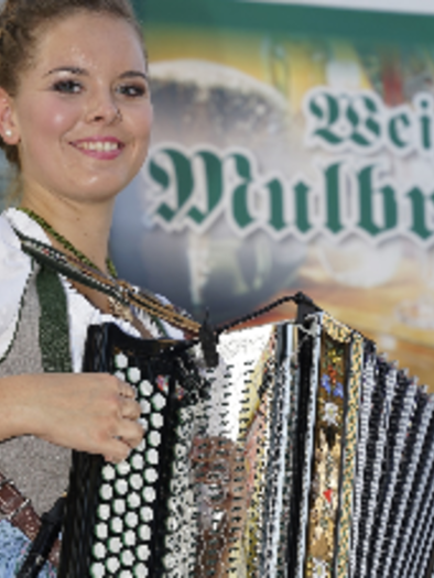 A cheerful young woman is wearing a traditional dress and playing an accordion. In the background, there is a banner that says "Weizer Mulbrat." | © KK