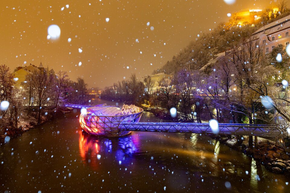 Eine verschneite Brücke, die über einen Fluss führt, ist bunt beleuchtet. Rundherum sind schneebedeckte Bäume und eine sanfte Winteratmosphäre. | © (c) Graz Tourismus - Harry Schiffer