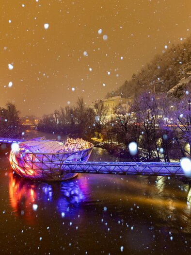A snowy bridge spanning a river is brightly lit. Surrounding it are snow-covered trees and a gentle winter atmosphere. | © (c) Graz Tourismus - Harry Schiffer
