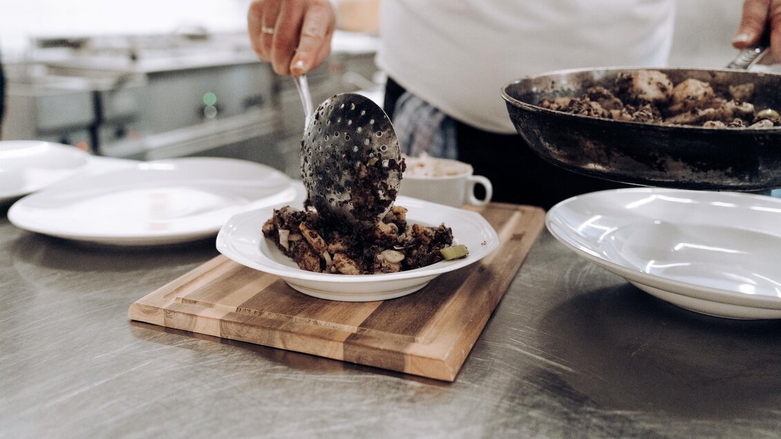 Ein Koch portioniert Essen auf einen Teller. Im Hintergrund sind weitere Teller und eine Küchenausstattung sichtbar. | © Martinhof