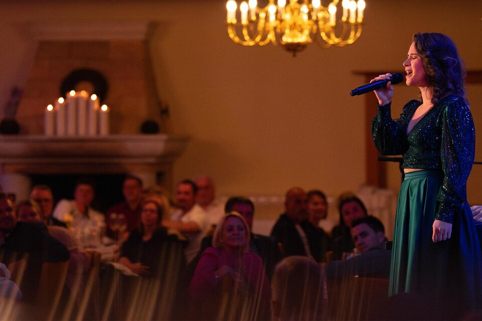 Eine Sängerin performt auf einer Bühne vor einem begeisterten Publikum. Im Hintergrund sind Kerzen und eine elegante Beleuchtung zu sehen.