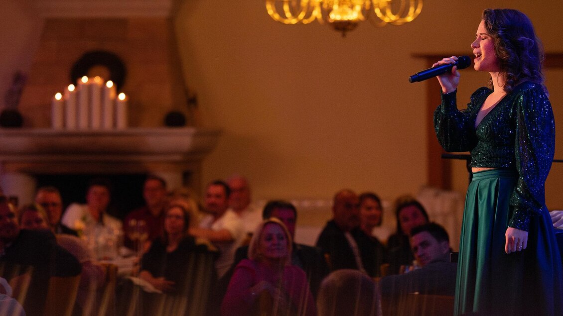 Eine Sängerin performt auf einer Bühne vor einem begeisterten Publikum. Im Hintergrund sind Kerzen und eine elegante Beleuchtung zu sehen.