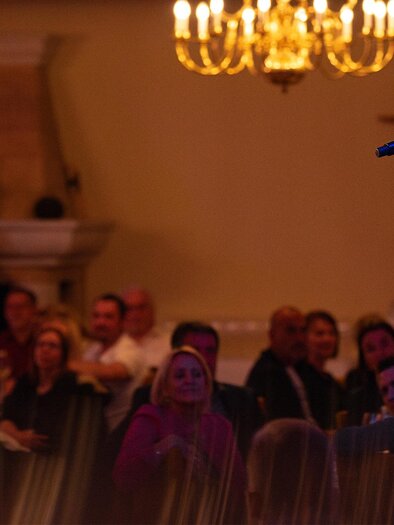 A singer performs on a stage in front of an enthusiastic audience. In the background, candles and elegant lighting are visible.