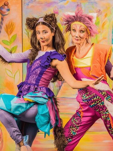 Two people in colorful costumes are posing in front of a colorful, painted background. They have joyful facial expressions and appear to be part of a performance. | © Theater HEUSCHRECK