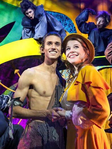 A colorful stage scene with performers in imaginative costumes. Two main characters are smiling at each other and standing in the foreground. | © Theater Liberi