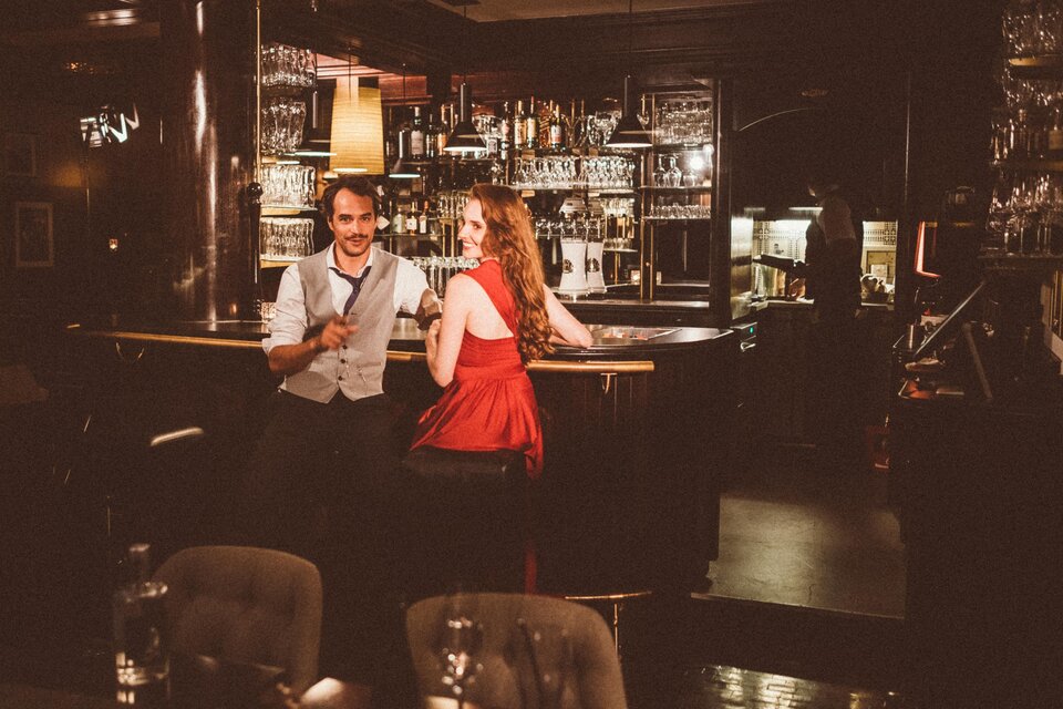 Ein Paar sitzt an der Bar in einem stilvollen Restaurant. Die Frau trägt ein rotes Kleid, während der Mann lächelt und einen Drink in der Hand hält. | © Thermenhotel Kowald