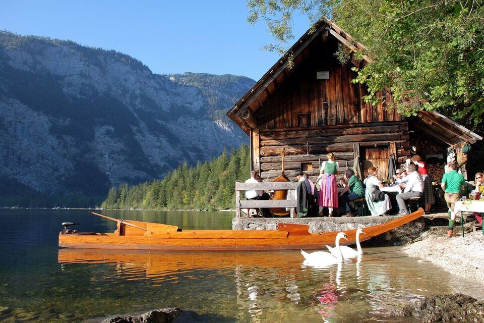 Ein malerisches Holzhaus am Ufer eines Sees, umgeben von Bergen und Bäumen. Gruppen von Menschen genießen die Natur, während Schwäne im Wasser schwimmen. | © Fremdenverkehrsverein Altaussee