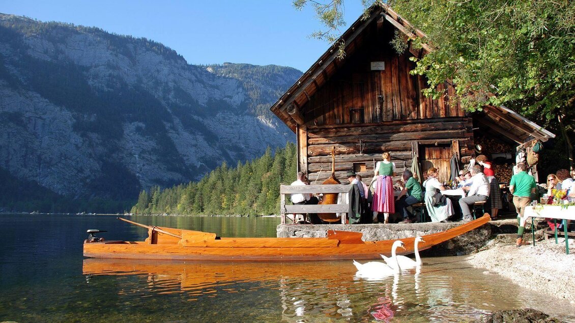 Ein malerisches Holzhaus am Ufer eines Sees, umgeben von Bergen und Bäumen. Gruppen von Menschen genießen die Natur, während Schwäne im Wasser schwimmen. | © Fremdenverkehrsverein Altaussee