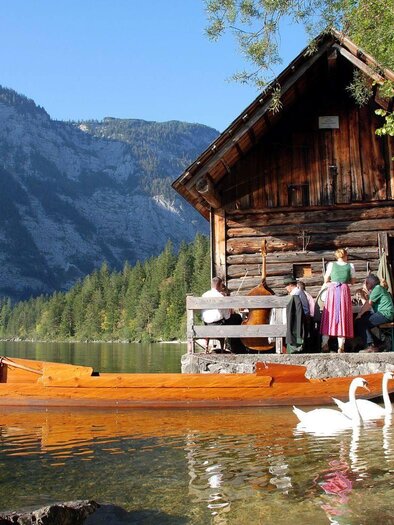 A picturesque wooden house by the shore of a lake, surrounded by mountains and trees. Groups of people enjoy nature while swans swim in the water. | © Fremdenverkehrsverein Altaussee