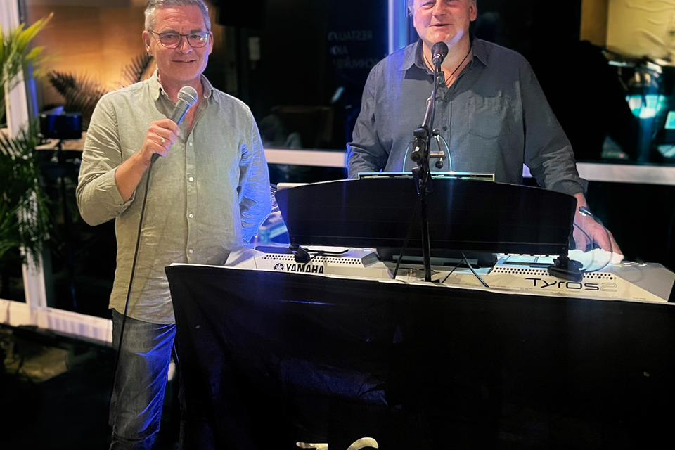 Zwei Männer stehen hinter einem Keyboard und Mikrofon und singen. Im Hintergrund sind Pflanzen und eine stilvolle Bar zu sehen. | © Das Kurhaus Bad Gleichenberg