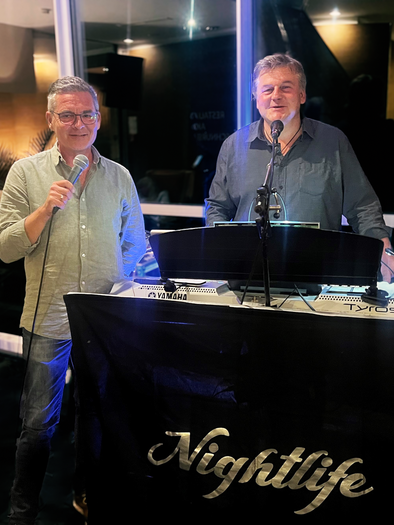 Zwei Männer stehen hinter einem Keyboard und Mikrofon und singen. Im Hintergrund sind Pflanzen und eine stilvolle Bar zu sehen. | © Das Kurhaus Bad Gleichenberg