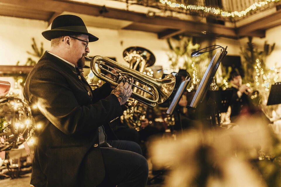Ein Musiker spielt Trompete in eleganter Abendkleidung. Im Hintergrund sind Lichter und andere Musiker zu sehen, die eine festliche Atmosphäre schaffen. | © Kunsthandwerk Schloss Burgau