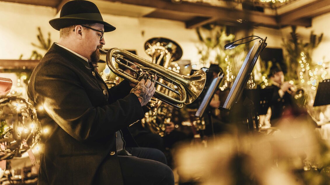 Ein Musiker spielt Trompete in eleganter Abendkleidung. Im Hintergrund sind Lichter und andere Musiker zu sehen, die eine festliche Atmosphäre schaffen. | © Kunsthandwerk Schloss Burgau