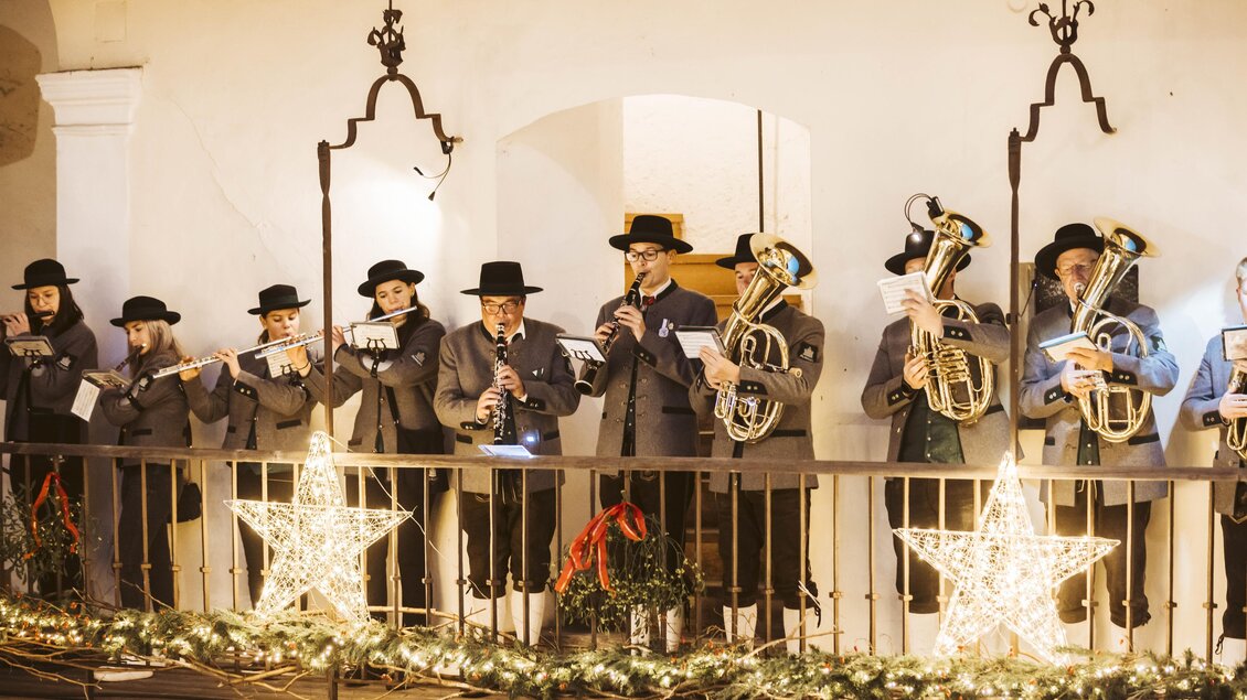 Eine Gruppe von Musikern spielt Instrumente auf einem Balkon. Die Szene ist festlich geschmückt mit Lichtern und Weihnachtssternen. | © Kunsthandwerk Schloss Burgau