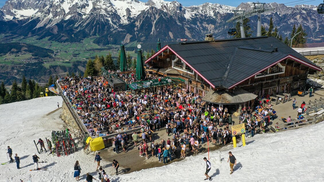 Ein belebtes Bergrestaurant voller Menschen vor einer beeindruckenden Alpenlandschaft. Schnee bedeckt den Boden und die Berge im Hintergrund sind mit Grün und Felsen durchzogen. | © Mario Egger