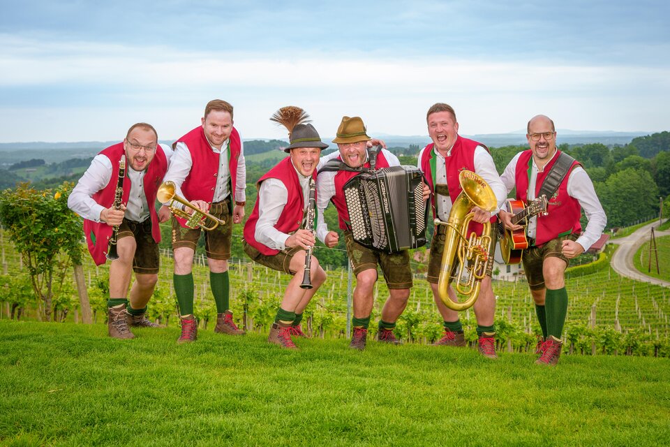 Eine Gruppe von sechs Männern in traditioneller bayerischer Tracht steht auf einer Wiese. Sie halten Musikinstrumente und lachen fröhlich in einer weingarten Umgebung. | © Markus Ponhold