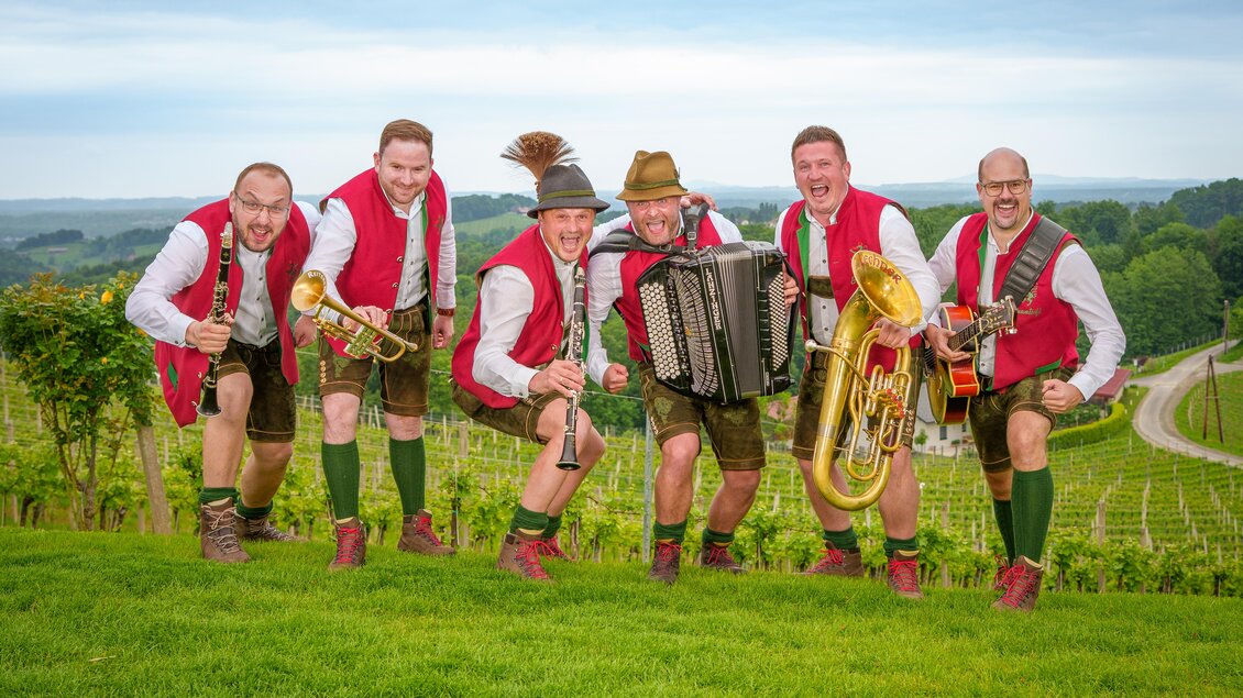Eine Gruppe von sechs Männern in traditioneller bayerischer Tracht steht auf einer Wiese. Sie halten Musikinstrumente und lachen fröhlich in einer weingarten Umgebung. | © Markus Ponhold
