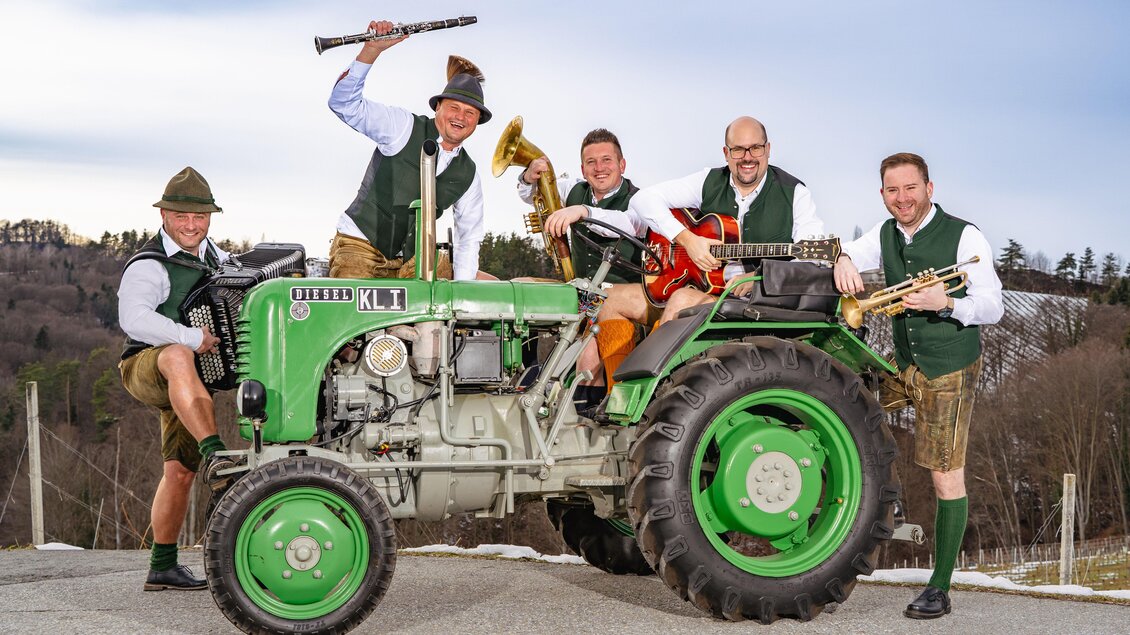 Eine Gruppe von fünf Männern in Trachten steht um einen grünen Traktor. Sie halten Musikinstrumente und posieren fröhlich vor einer ländlichen Landschaft. | © Markus Ponhold