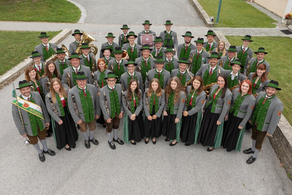 Eine Gruppe von Menschen steht in traditioneller Tracht in einem Außenbereich. Sie lächeln und posieren gemeinsam für das Foto. | © Trachtenmusikkapelle St. Magdalena a. L.