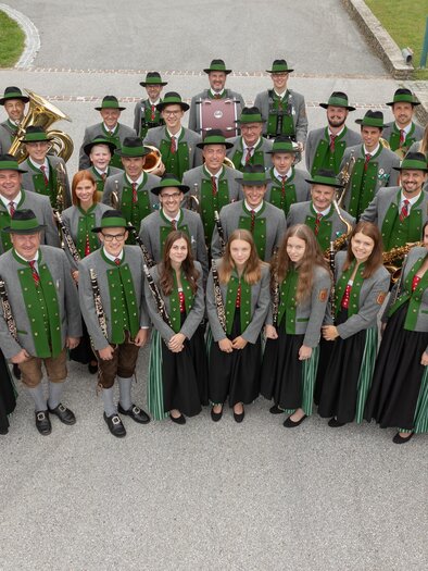 Eine Gruppe von Menschen steht in traditioneller Tracht in einem Außenbereich. Sie lächeln und posieren gemeinsam für das Foto. | © Trachtenmusikkapelle St. Magdalena a. L.