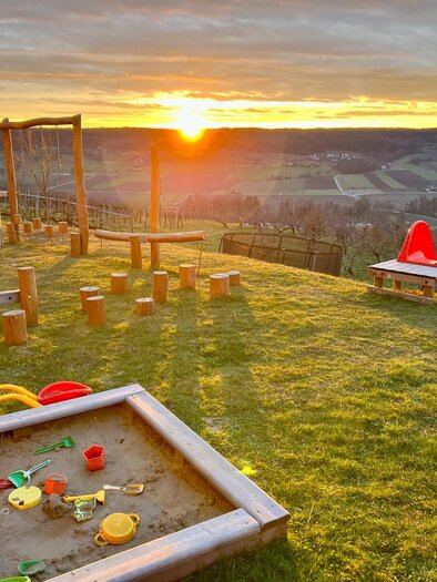 A playground with a sandpit, a slide, and climbing frames. In the background, one can see a beautiful sunset over hills. | © Markus Lugitsch