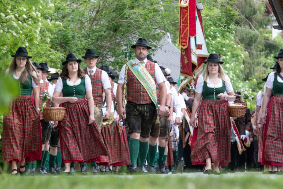Eine Gruppe von Menschen in traditioneller bayerischer Tracht marschiert durch eine grüne Umgebung. Einige tragen Körbe, während andere Fahnen präsentieren. | © Trachtenkapelle Donnersbach