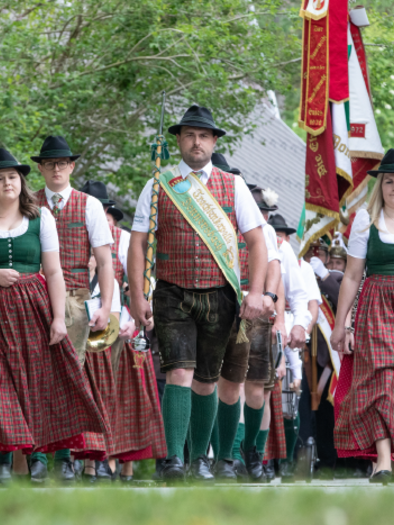 A group of people in traditional Bavarian attire marches through a green environment. Some are carrying baskets while others are presenting flags. | © Trachtenkapelle Donnersbach