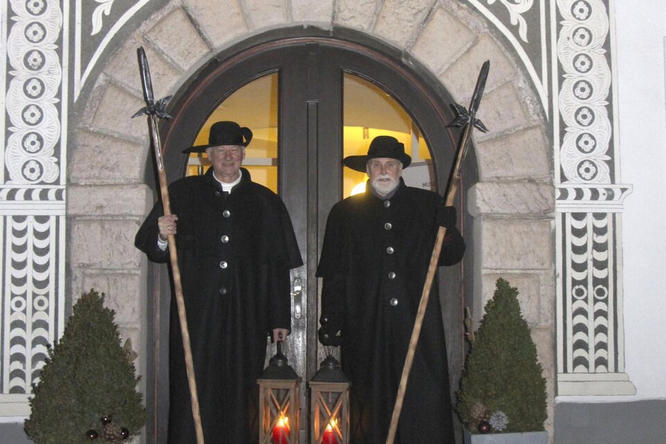 Zwei Männer in traditionellen schwarzen Anzügen stehen vor einem historischen Gebäude. Sie halten lange Stäbe und es sind Laternen zu sehen. | © Stadtgemeinde Eisenerz