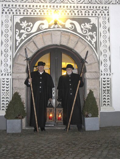 Two men in traditional black suits stand in front of a historic building. They are holding long poles and lanterns can be seen. | © Stadtgemeinde Eisenerz