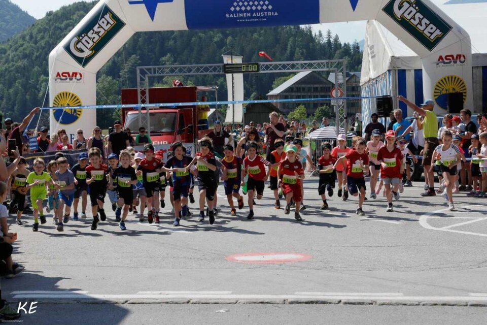 Eine Gruppe von Kindern läuft energisch über die Ziellinie eines Wettkampfs. Im Hintergrund sind Berge und Zuschauern zu sehen, die die Veranstaltung feiern. | © Kainzinger Erich