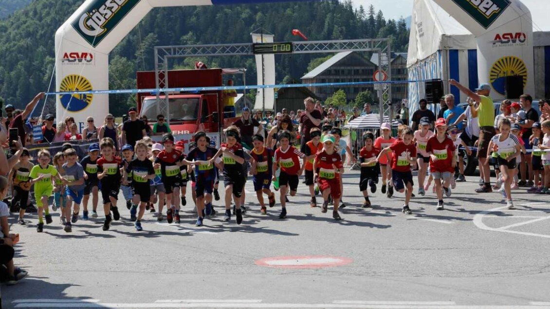 Eine Gruppe von Kindern läuft energisch über die Ziellinie eines Wettkampfs. Im Hintergrund sind Berge und Zuschauern zu sehen, die die Veranstaltung feiern. | © Kainzinger Erich