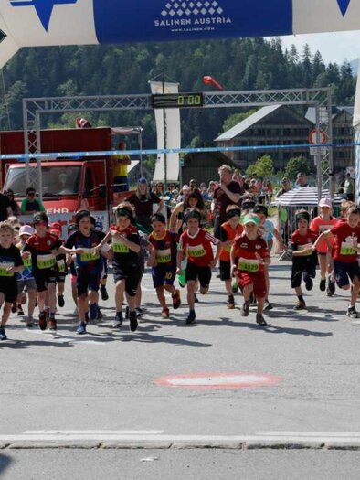 A group of children runs energetically across the finish line of a competition. In the background, mountains and spectators can be seen celebrating the event. | © Kainzinger Erich