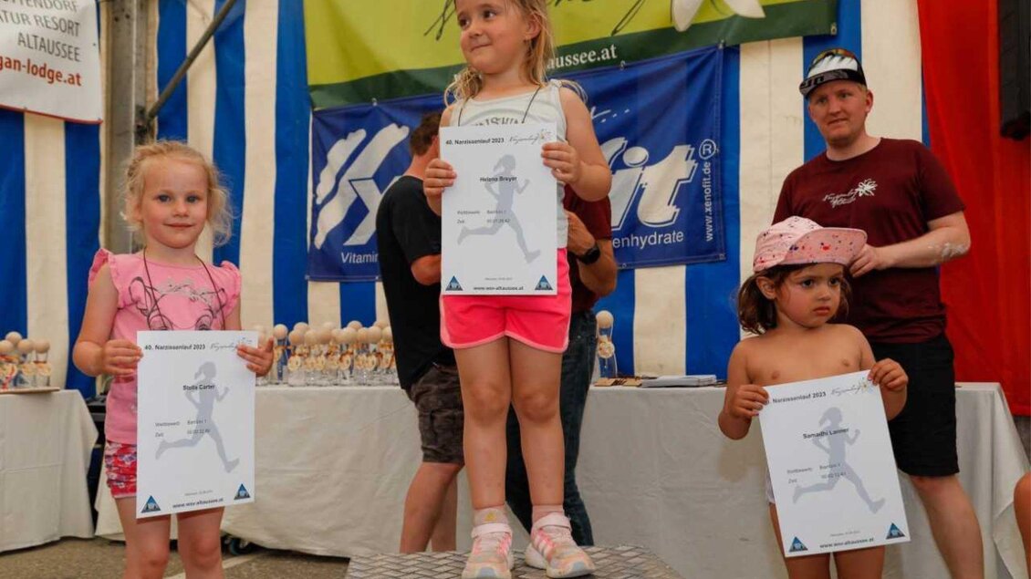 Drei Kinder stehen auf dem Siegertreppchen und halten Urkunden in den Händen. Im Hintergrund sind bunte Zeltwände und eine Trophäensammlung zu sehen. | © Kainzinger Erich