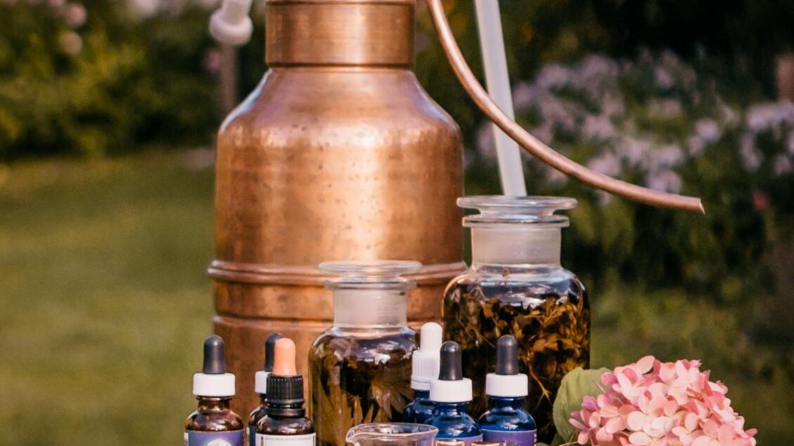 Eine Kupferdestille steht auf einem Tisch im Freien, umgeben von verschiedenen Glasfläschchen mit Kräuterextrakten. Auf dem Tisch liegen auch einige Blumen und Dekorative Elemente. | © Petra Stückelschwaiger