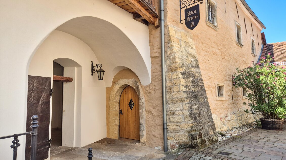 Ein historisches Gebäude mit einem schönen Eingang und einer hölzernen Tür. Der Eingang ist von einer Steinfassade umgeben, und es gibt eine Laterne und Pflanzen im Bereich. | © @Winkler-Hermaden