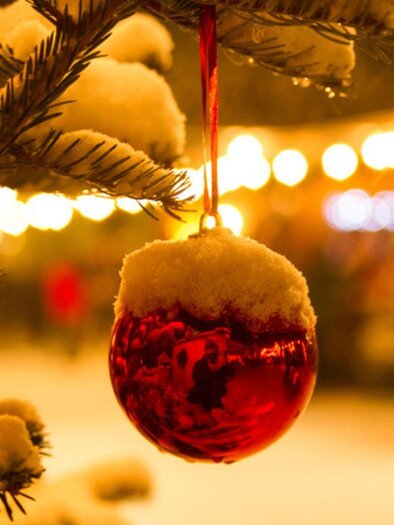 A red Christmas ornament hangs on a snow-covered tree. In the background, festive lights of a Christmas market can be seen. | © Silvia Rastl