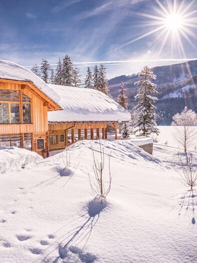 Ein charmantes Holzhaus im Schnee, umgeben von einer winterlichen Landschaft. Die Sonne strahlt über die Bergkulisse und sorgt für eine traumhafte Atmosphäre. | © Kohlröserlhütte Genuss am See
